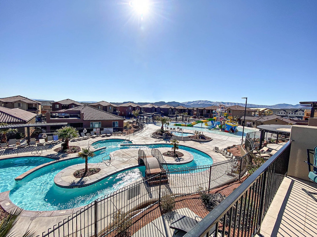 A scenic view displays a winding pool surrounded by palm trees, with vibrant water slides visible nearby. The sun shines brightly in a clear blue sky, illuminating the resort-style setting. Cozy lounge chairs are arranged along the pool deck.