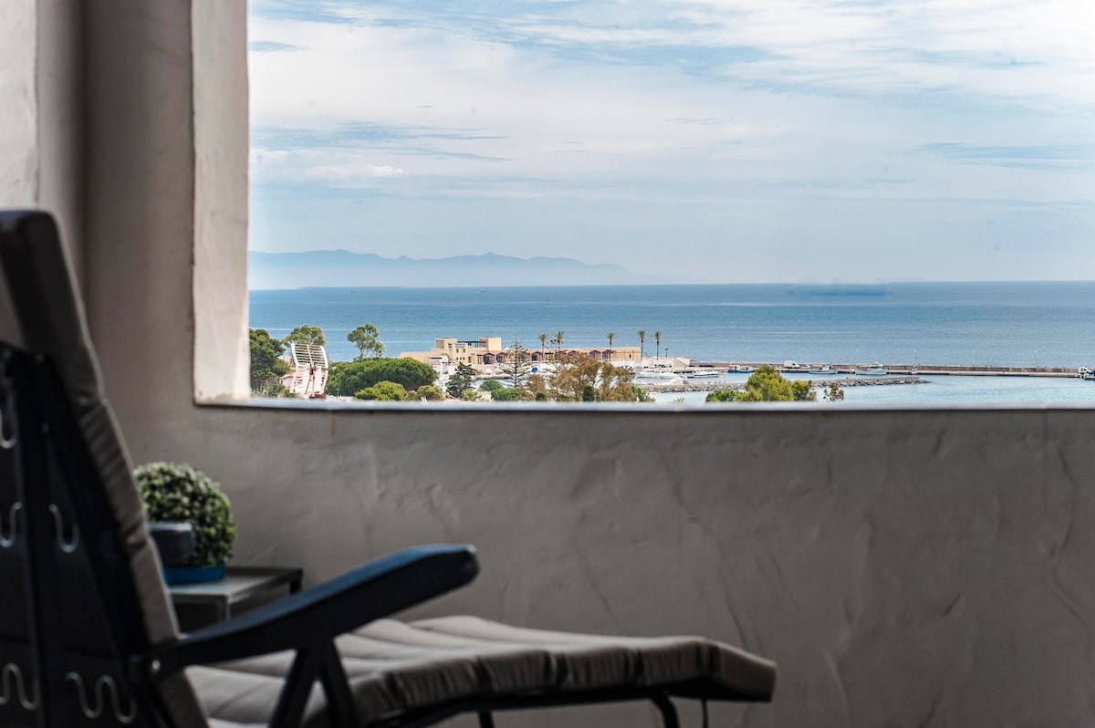 A large window frame offers a scenic view of the Mediterranean Sea and a nearby marina. A single chair with a soft cushion is positioned nearby, while greenery is placed on a small side table. The tranquil water contrasts with distant landforms on the horizon.