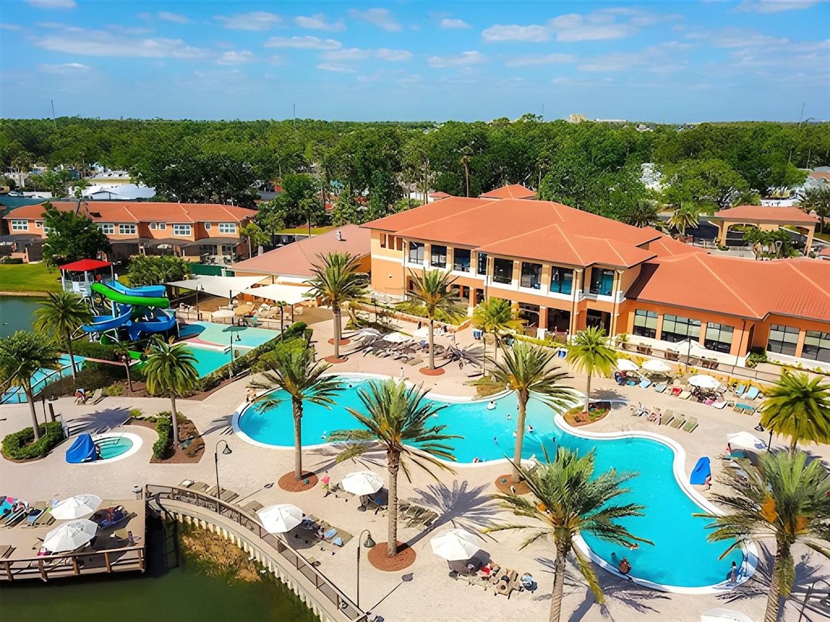 The resort features a large pool area surrounded by palm trees, with lounge chairs scattered along the water's edge. A clubhouse is visible in the background, and colorful water slides are positioned nearby, inviting guests to enjoy the aquatic amenities.