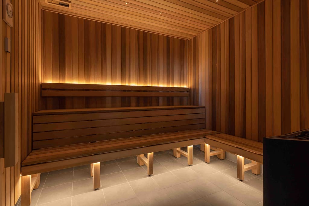 A sauna room features warm wooden paneling and soft ambient lighting, providing a serene environment for relaxation. Wooden benches line the room, arranged to accommodate multiple guests. The floor is tiled, and the space is designed to enhance a cozy atmosphere.