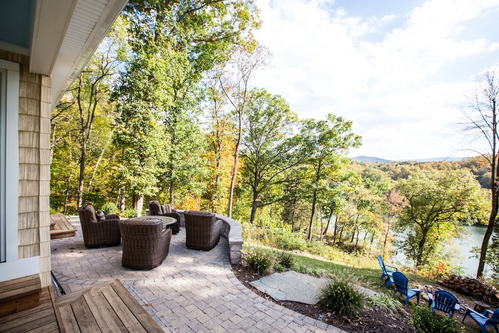 A spacious patio area is surrounded by lush trees, offering seating on woven armchairs. A stone pathway leads towards the river, where scenic views of the landscape are visible. Two blue chairs are placed nearby, providing additional outdoor seating options.