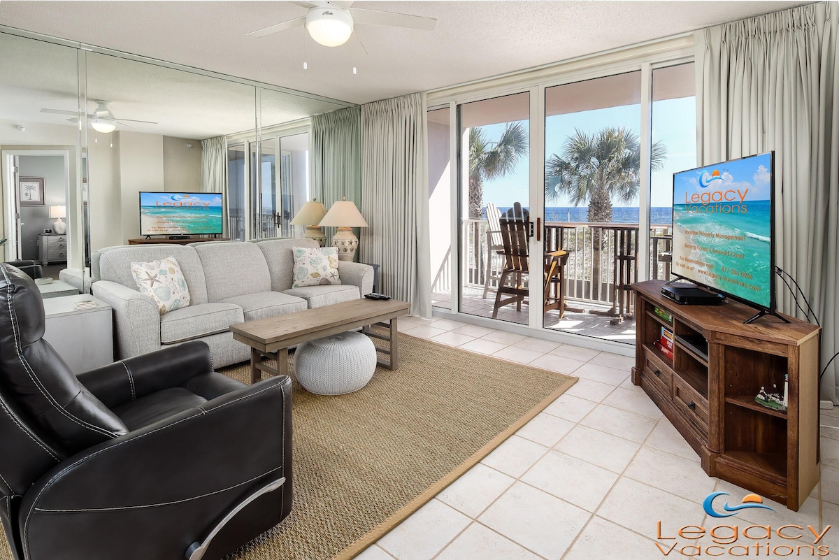 The living room showcases a light-colored sofa and a black reclining chair, with a wooden coffee table positioned between them. A large TV is mounted on a cabinet against a mirrored wall. Floor-to-ceiling doors lead to a private balcony overlooking palm trees and the Gulf.