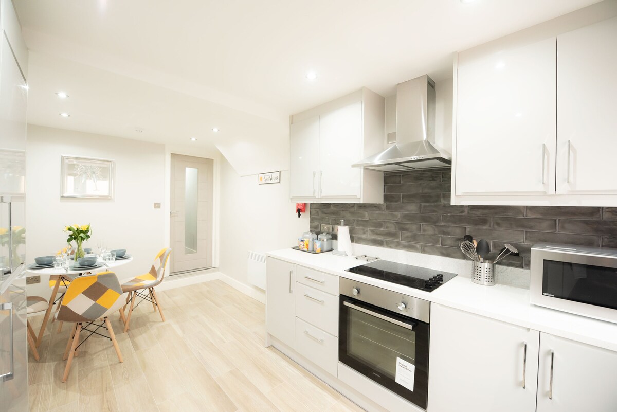 A modern kitchen features sleek cabinetry and stainless steel appliances, including a stove and microwave. A dining table with colorful chairs is positioned nearby, illuminated by warm overhead lights. The space is well-organized and exudes a clean, contemporary design.