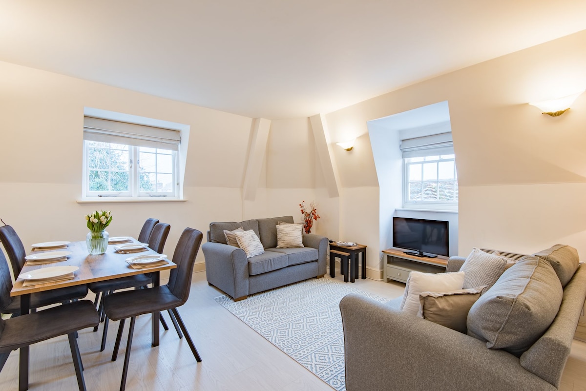 A spacious sitting and dining area features two comfortable sofas, a flat-screen TV, and a dining table set for six. Natural light fills the room through large windows, illuminating the neutral color palette and a light area rug beneath the furniture.
