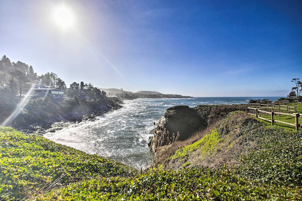 A scenic view of the coastline is presented, featuring rolling waves and rocky outcrops under a bright blue sky. Lush greenery borders the shoreline, while sunlight illuminates the landscape, highlighting the contours of the land and water.
