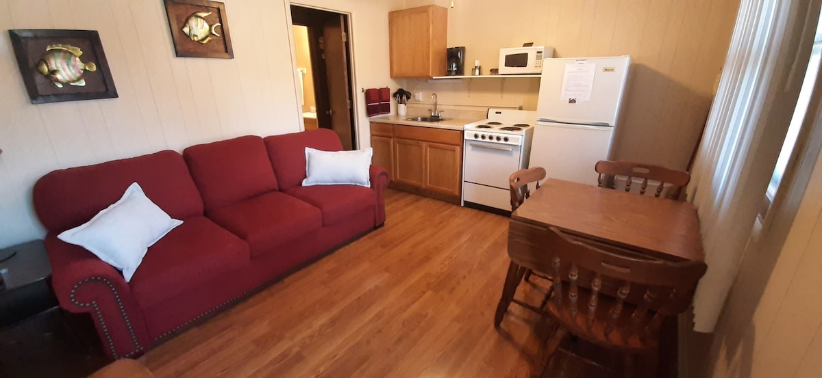 A cozy living area features a red couch with white pillows, a wooden dining table with seating for four, and a well-equipped kitchen area. A refrigerator, stove, and microwave are visible, along with wooden cabinets and fish art adorning the walls.
