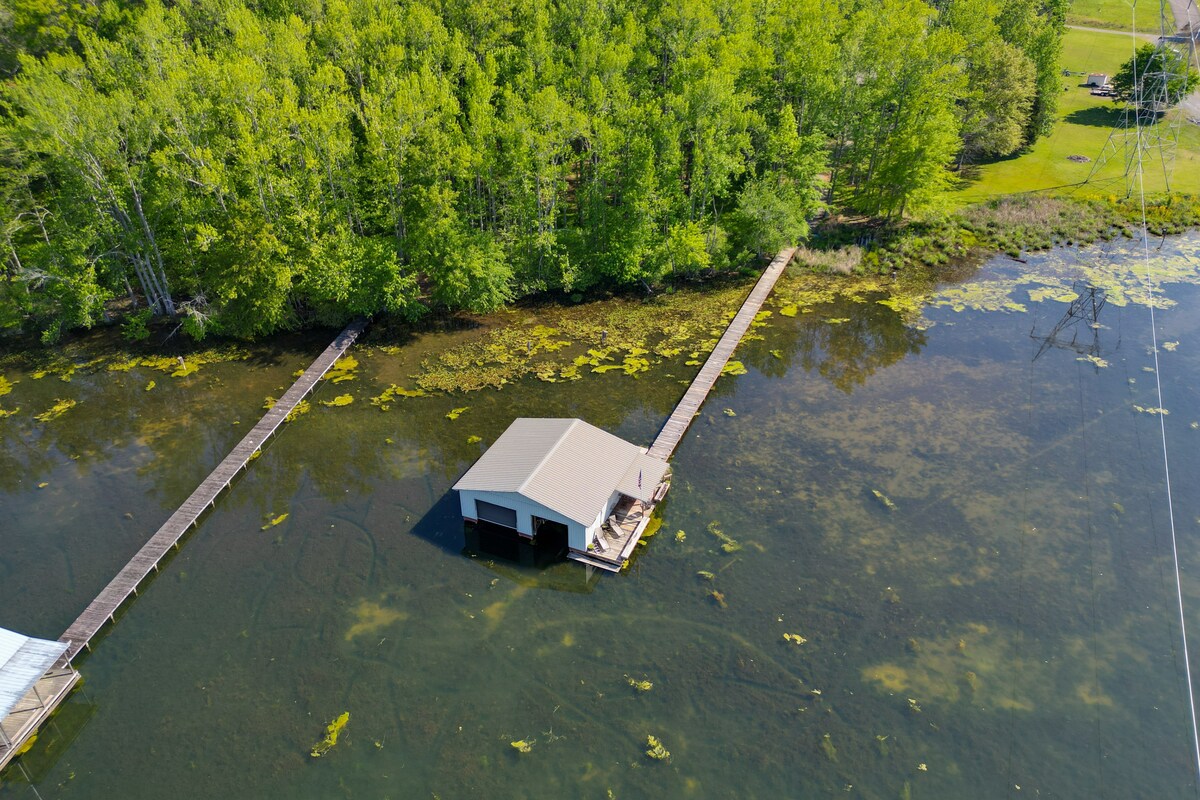 An aerial view captures a boathouse situated on Guntersville Lake, surrounded by lush greenery. The photograph showcases wooden piers extending towards the water, providing access to the lakeside, with patches of lily pads visible in the water.