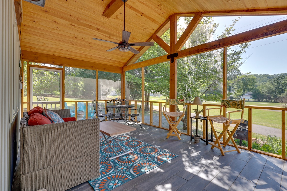 A screened-in porch features a ceiling fan and wood paneling, furnished with a woven couch, a hammock chair, and two small tables. A colorful area rug adds vibrancy, while large windows provide views of the surrounding greenery.
