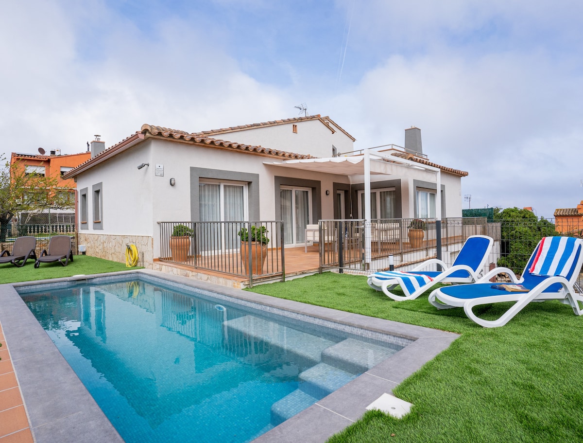 A comfortable house features a small private pool surrounded by a well-maintained lawn. Loungers with blue and white stripes sit near the pool, offering a relaxing area to enjoy the outdoors. The patio includes access to the living spaces through large glass doors.