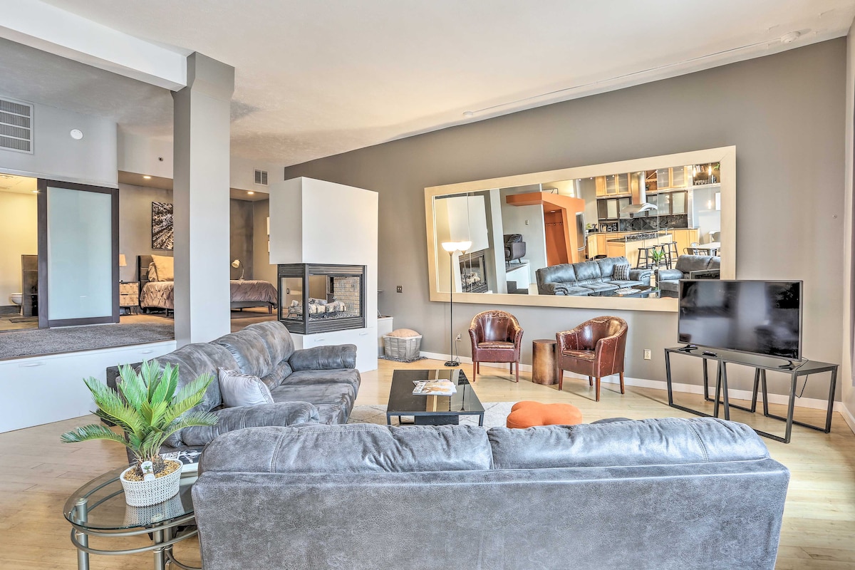 A spacious living area features a cozy seating arrangement with a plush gray sofa and two brown armchairs. A large mirror reflects the room's open layout, showcasing a decorative fireplace and flat-screen TV. The modern kitchen area is visible in the background.