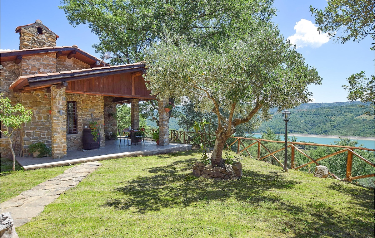 The charming exterior of a stone vacation home is seen, framed by lush olive trees. A covered patio offers seating with views of the lake in the distance. A stone pathway leads through the well-maintained garden, enhancing the serene outdoor atmosphere.