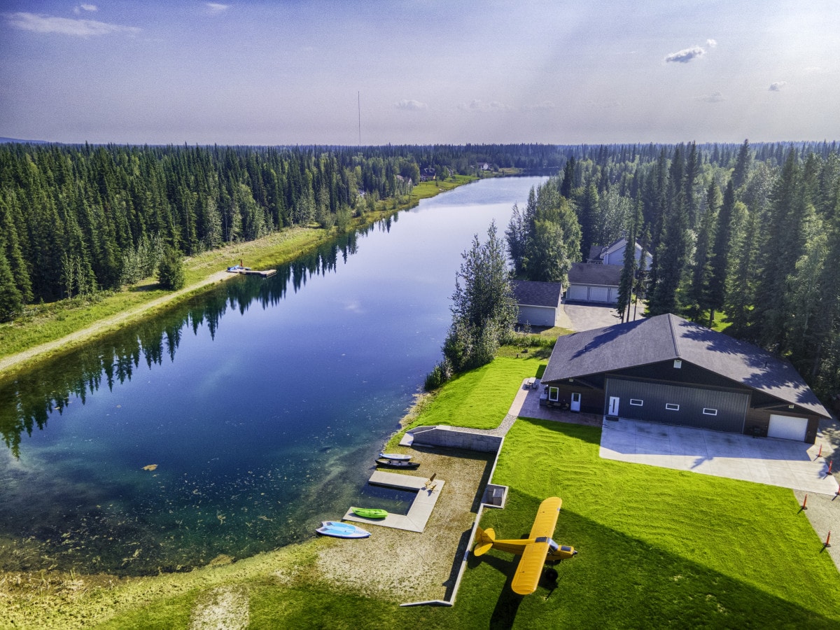 An aerial view of a serene lakeside property is captured, showcasing a lush green lawn leading to the calm water. A vibrant yellow airplane sits nearby, and paddleboards are visible on the concrete dock along the shoreline, framed by dense trees and a clear sky.