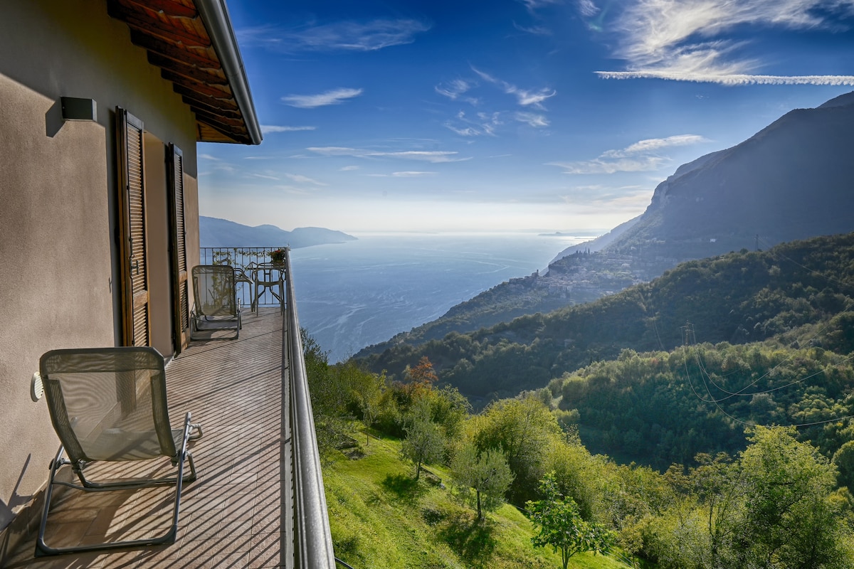 A wooden balcony offers a stunning view of the lake and surrounding mountains. Two lounge chairs are placed on the balcony, providing a comfortable space to enjoy the panoramic scenery. The lush greenery slopes gently down towards the water, with a clear sky above.