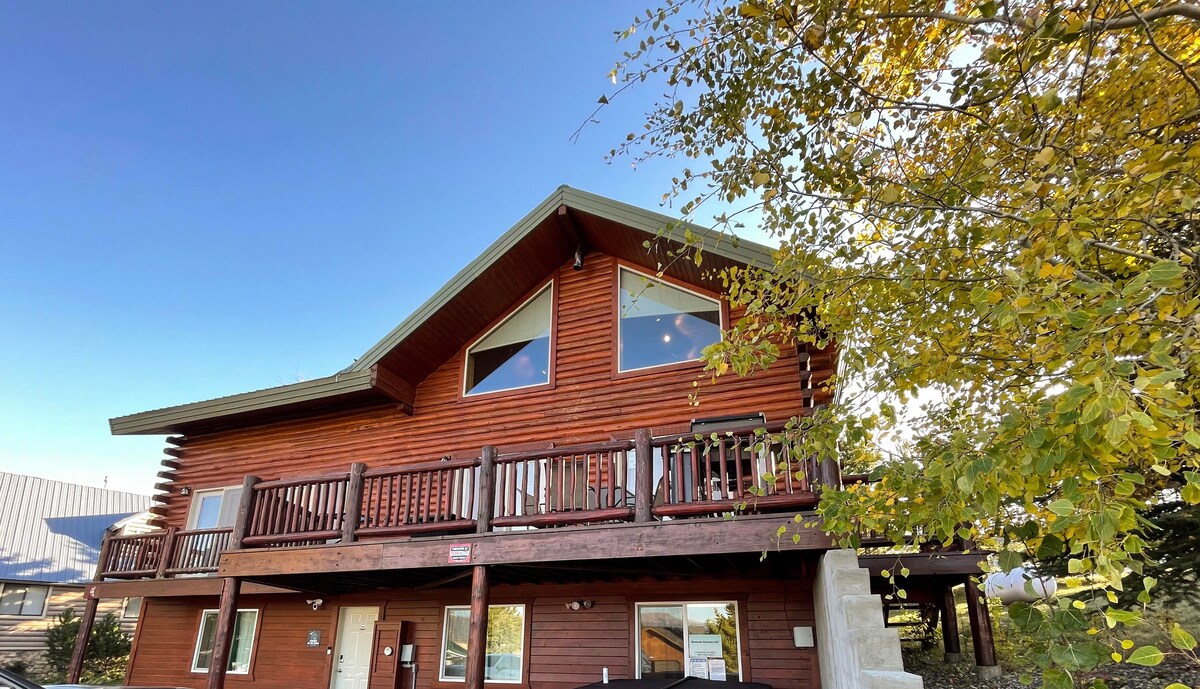 A spacious cabin is shown, constructed from natural wood with a sloped roof. Large triangular windows allow natural light to fill the interior. A wooden balcony extends along the front, adorned with sturdy railings. Green foliage frames the cabin, highlighting its natural surroundings.