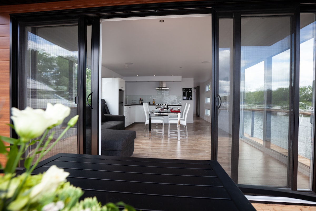 An open layout is showcased through large sliding glass doors, leading to a modern kitchen and dining area. Light wooden flooring complements the sleek furnishings. A comfortable seating area is visible, along with a dining table set for four, creating an inviting space for guests.