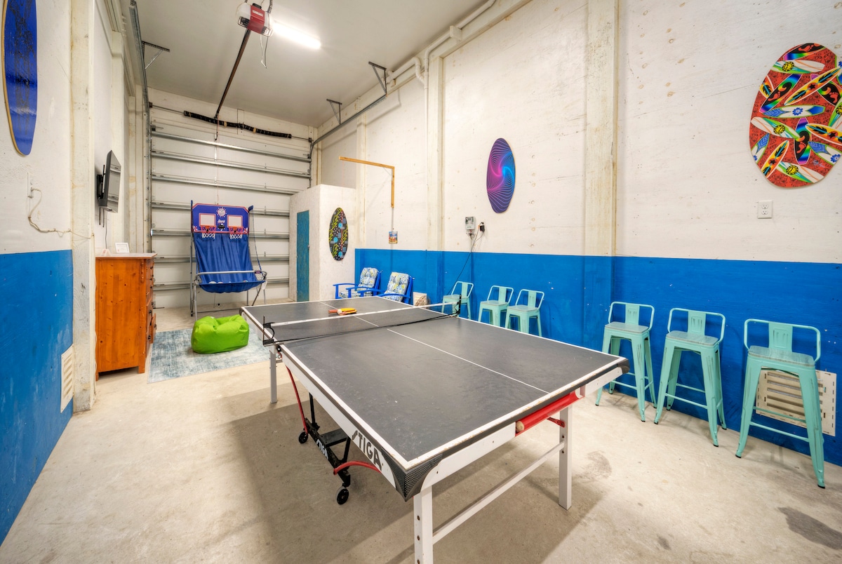 A game room is set up in a garage space, featuring a ping pong table at the center. Colorful wall art enhances the environment, while metal chairs and a bean bag provide additional seating. The garage door remains open, allowing natural light to enter.
