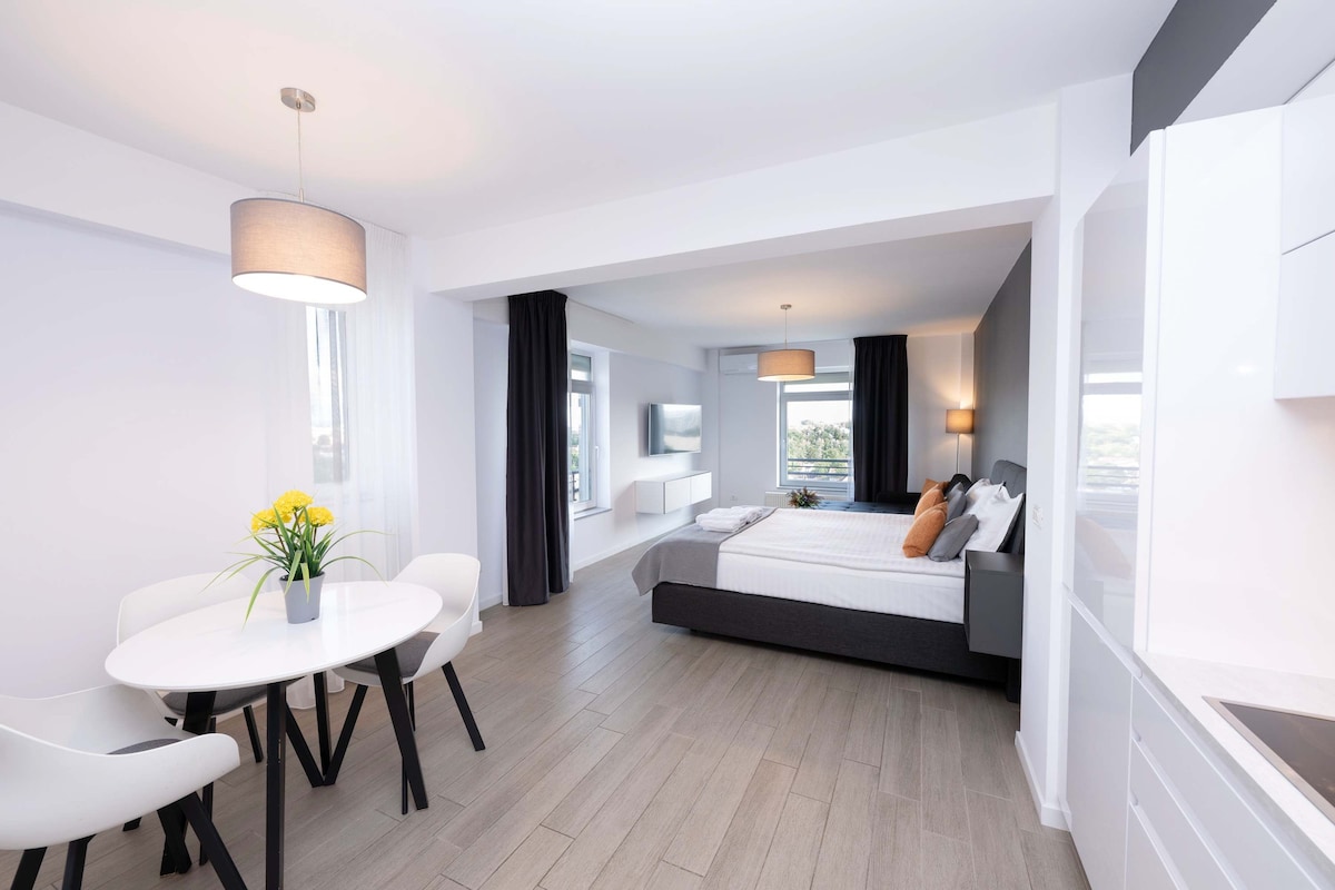 A modern studio layout features a spacious sleeping area with a king size bed and a sofa bed. A small dining table with four white chairs is positioned nearby. Large windows allow ample natural light, enhancing the airy feel of the room.