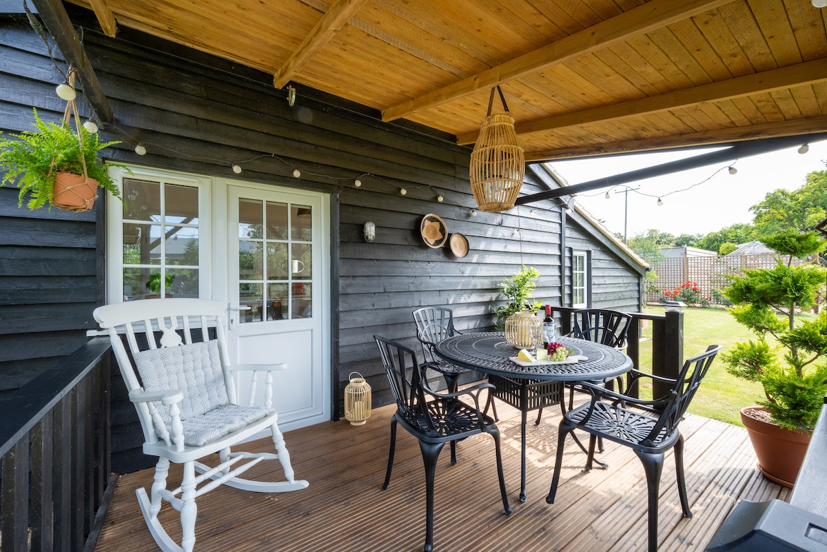 A covered deck area features a round table surrounded by several black chairs. A white rocking chair is positioned nearby. Decorative items and plants adorn the walls, and soft lighting is provided by string lights overhead, creating a comfortable outdoor space.