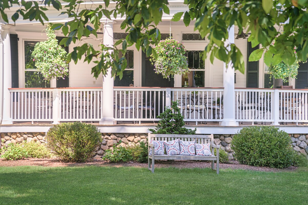 A welcoming porch is framed by lush greenery, featuring a wooden bench adorned with colorful patterned cushions. Hanging planters add a touch of vibrancy, while the stone foundation complements the white railing and columns of the house.