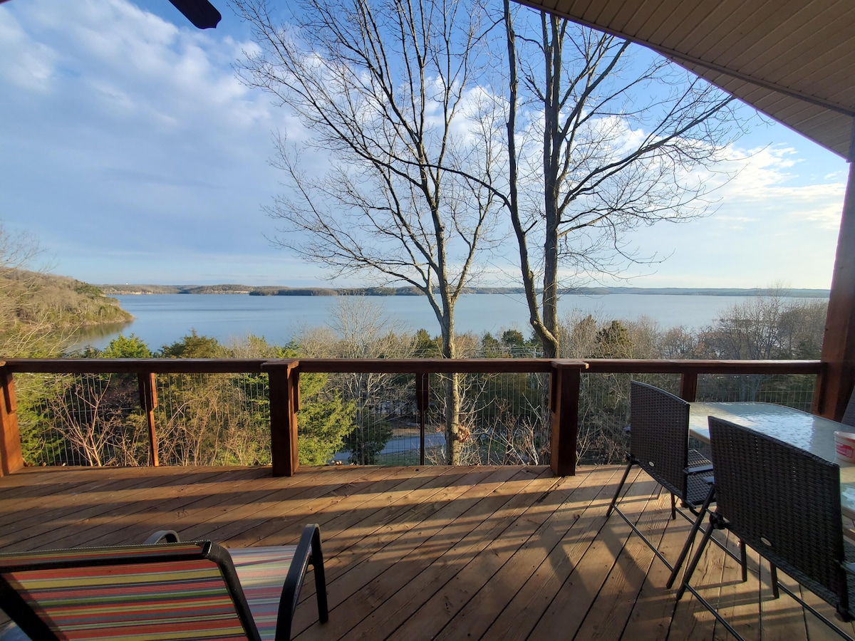 A spacious deck overlooks expansive views of Beaver Lake, framed by trees and open sky. Nearby seating areas are positioned for comfort, inviting guests to relax while enjoying the waterfront scenery.