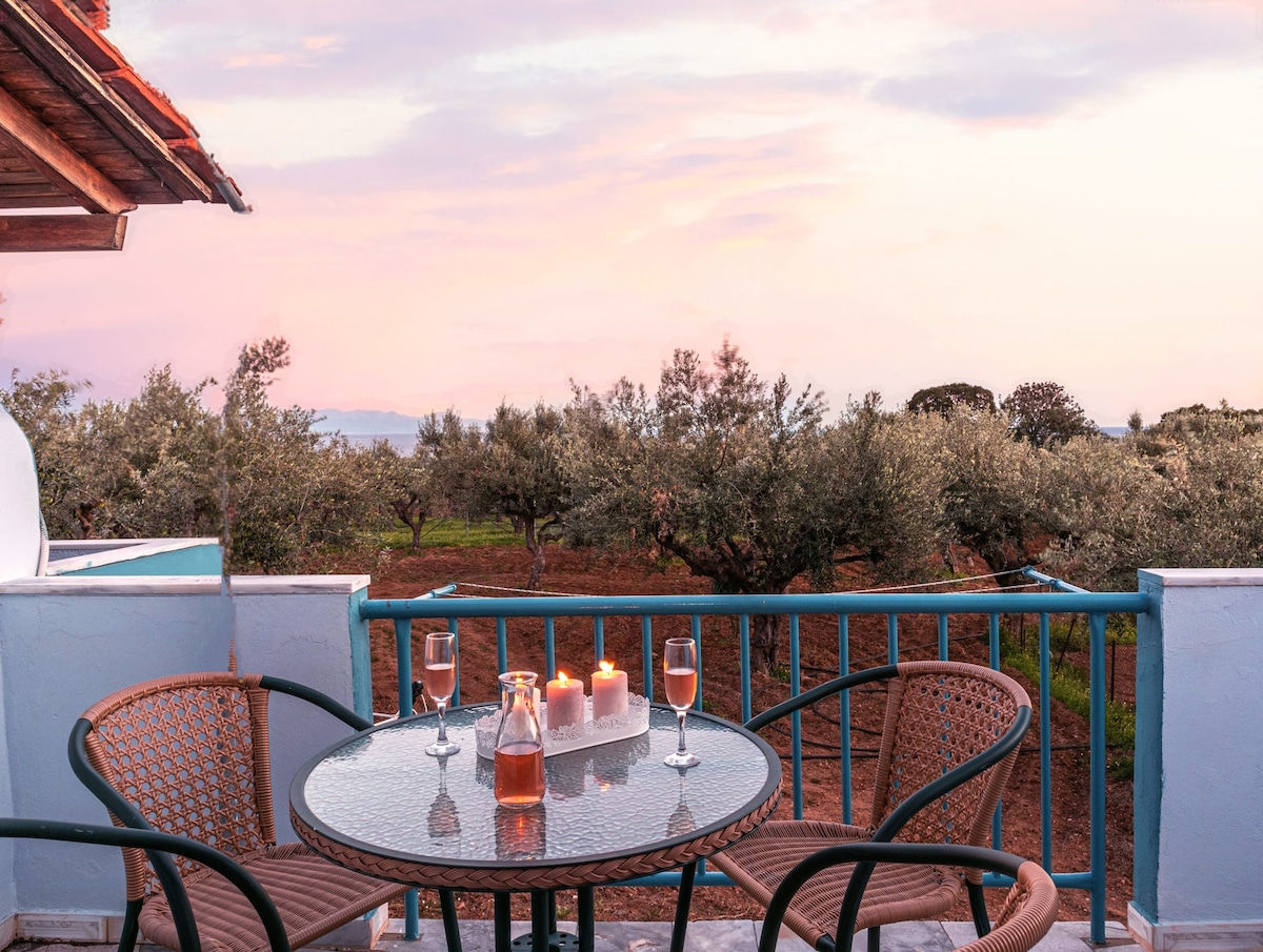 A relaxing balcony features a round glass table surrounded by two chairs. A glass pitcher and two glasses stand on the table, accompanied by glowing candles. The view showcases an olive grove with a soft pink sky at sunset in the background.