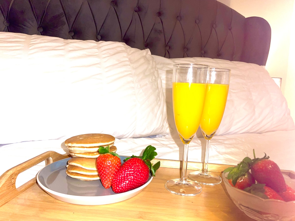 On a wooden tray, a plate contains stacked pancakes alongside fresh strawberries. Two glasses filled with orange juice sit beside the plate, in front of a neatly arranged bed featuring a tufted headboard and crisp white linens.