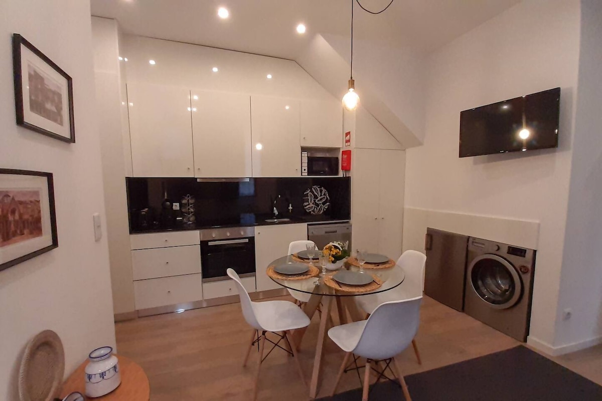 A modern kitchen and dining area is presented, featuring sleek white cabinetry and a glass dining table surrounded by four white chairs. Appliances such as a microwave and oven are integrated into the design, with a washing machine visible on one side, under warm ceiling lights.