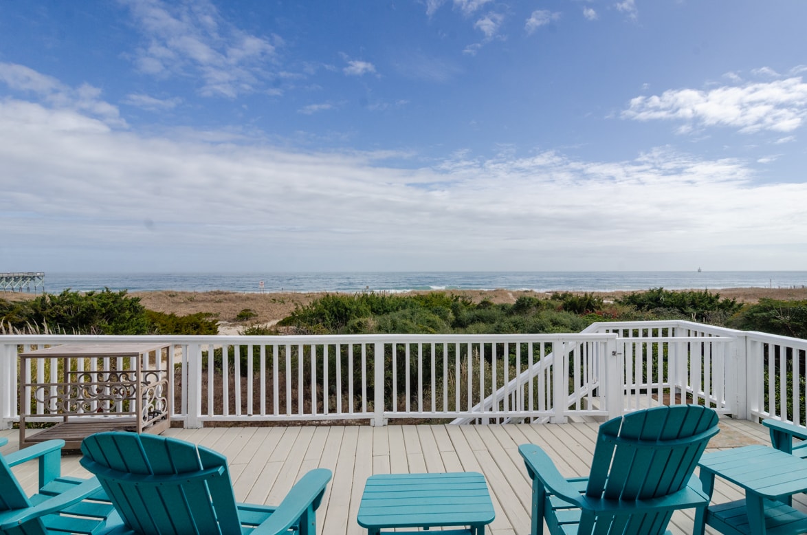 An expansive oceanfront deck is featured, furnished with several blue Adirondack chairs facing the Atlantic Ocean. The deck overlooks a sandy beach and lush green vegetation that borders the shoreline, with gentle waves visible under a clear blue sky.
