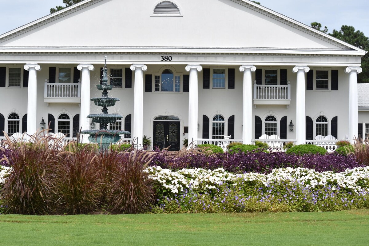 A grand white building is framed by tall columns and adorned with decorative black doors and windows. A fountain is positioned in the foreground, surrounded by vibrant flower beds featuring a mix of purple and white blooms, set against a lush green lawn.