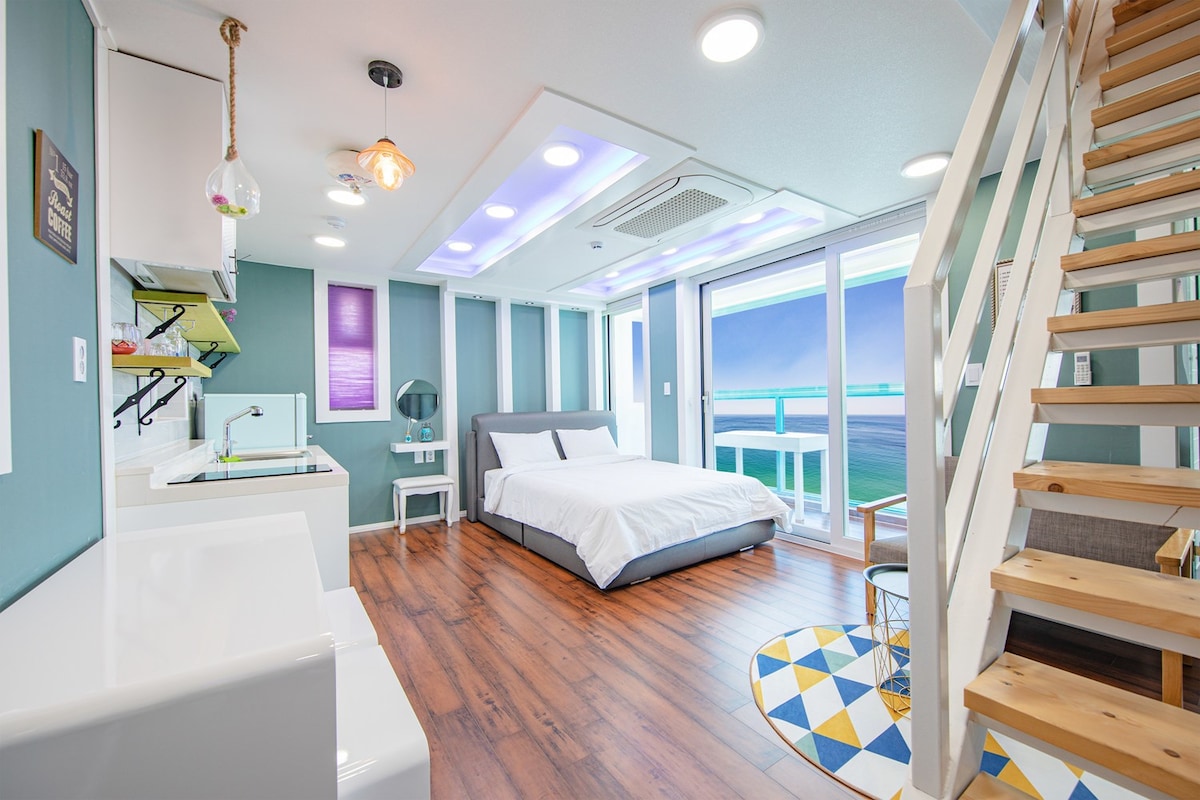 A bright and airy living space features a spacious bed beside a large window with stunning ocean views. A stylish staircase leads to a lofted area. Modern kitchen amenities are present, enhanced by contemporary decor and a decorative area rug.