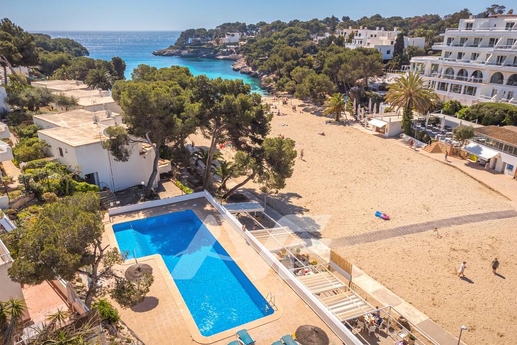 An aerial view captures a swimming pool surrounded by sun loungers, overlooking a sandy beach that meets clear blue waters. Lush greenery and palm trees provide shade, enhancing the inviting coastal atmosphere. Residential buildings are visible in the background, merging natural and architectural elements.