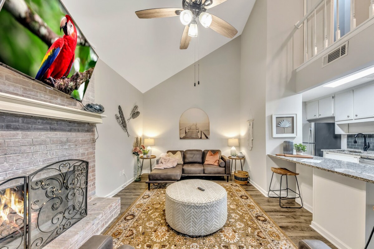 A cozy living area is highlighted by a textured area rug and a spacious circular ottoman at the center. A comfortable sofa is placed against a decorative stone fireplace, with a large TV mounted above. Soft lighting enhances the welcoming ambiance, complemented by ceiling fans overhead.