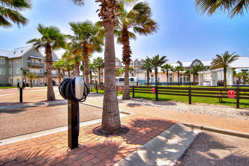 A landscaped area features several palm trees lining a pathway. Charging stations for electric vehicles are positioned on the left, with a grassy area visible in the background. The vibrant blue sky enhances the inviting atmosphere of the outdoor space.
