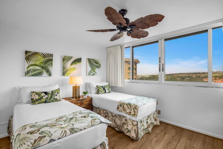 Camas individuales vestidas con ropa de cama tropical, enmarcadas por obras de arte de hojas de palmera y una amplia vista a la ventana.