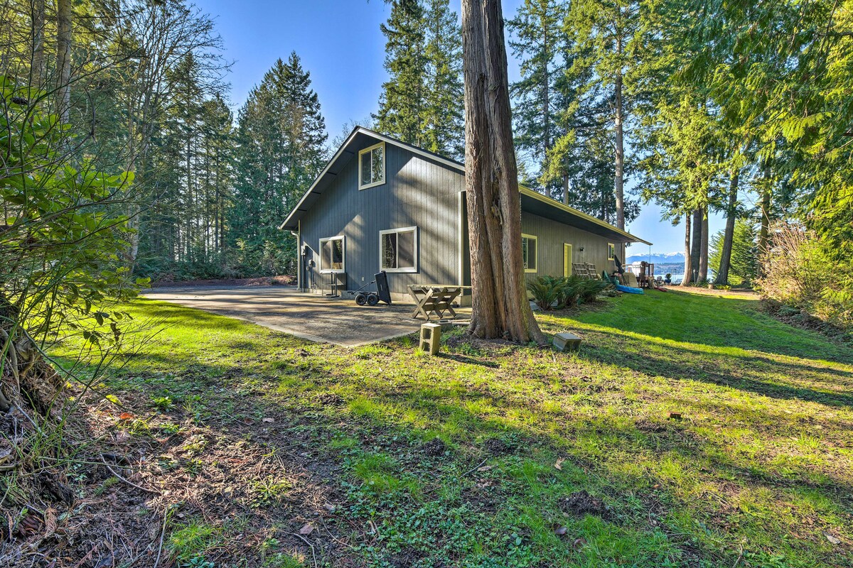 The cabin is nestled among tall evergreens, providing a serene outdoor setting. The exterior features a sleek grey facade, large windows, and a spacious driveway. A picnic table is visible, inviting outdoor gatherings. The distant shoreline hints at nearby waterfront access.