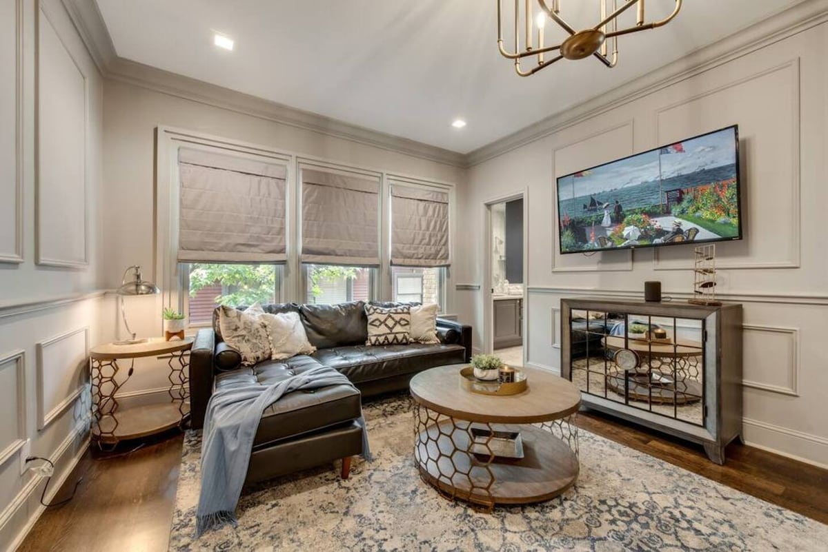 A stylish living room is showcased, featuring a dark leather sofa complemented by patterned throw pillows. A round wooden coffee table rests on a decorative area rug. Large windows with roman shades allow for natural light, while a modern chandelier and a television enhance the space.