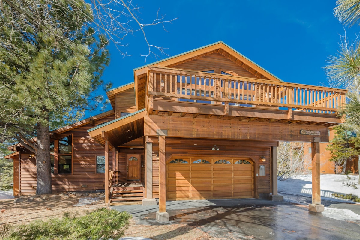 The exterior of the wooden house features multiple levels and a spacious front deck. Large windows reflect natural light, while a covered parking area shows two garage doors. Surrounding trees provide shade, and a path leads to the entrance, set against a clear blue sky.