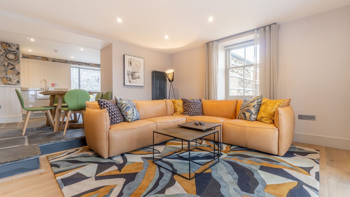 A spacious living area is presented with a large, deep-cushioned corner sofa featuring various textured cushions. A modern coffee table sits at the center on a patterned area rug, while natural light enters through a nearby window complemented by soft curtains.