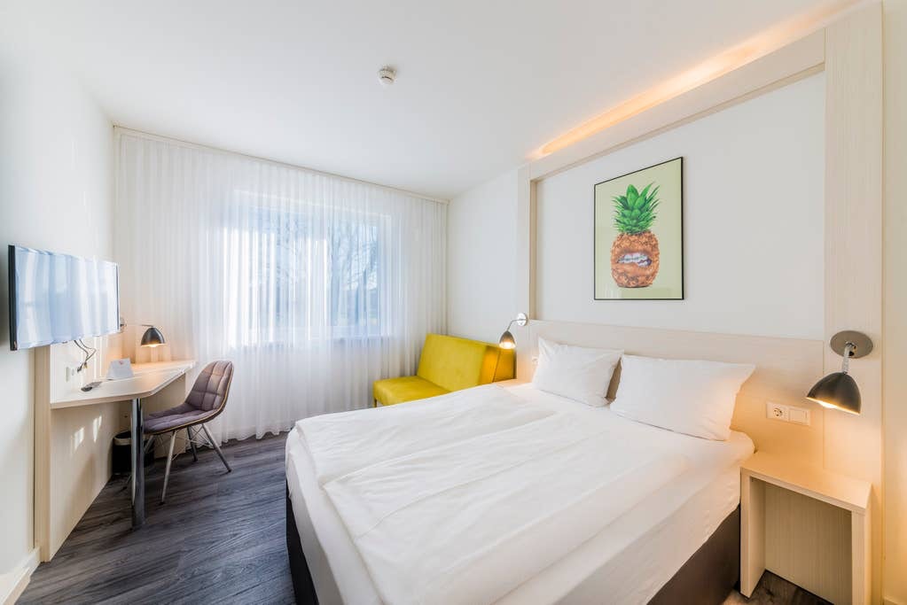 A light-filled room features a spacious bed with crisp white linens and decorative pillows. A small desk with a chair is positioned near a window. A yellow seating area adds a pop of color, while a framed art piece of a pineapple decorates the wall.