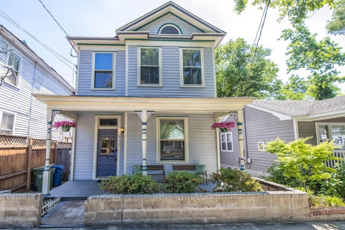 A charming Victorian-style duplex with a light blue exterior is framed by lush greenery. The front porch features welcoming seating beneath colorful flower boxes, inviting relaxation. Sunlight filters through the trees, highlighting the character of this downtown Wilmington home.