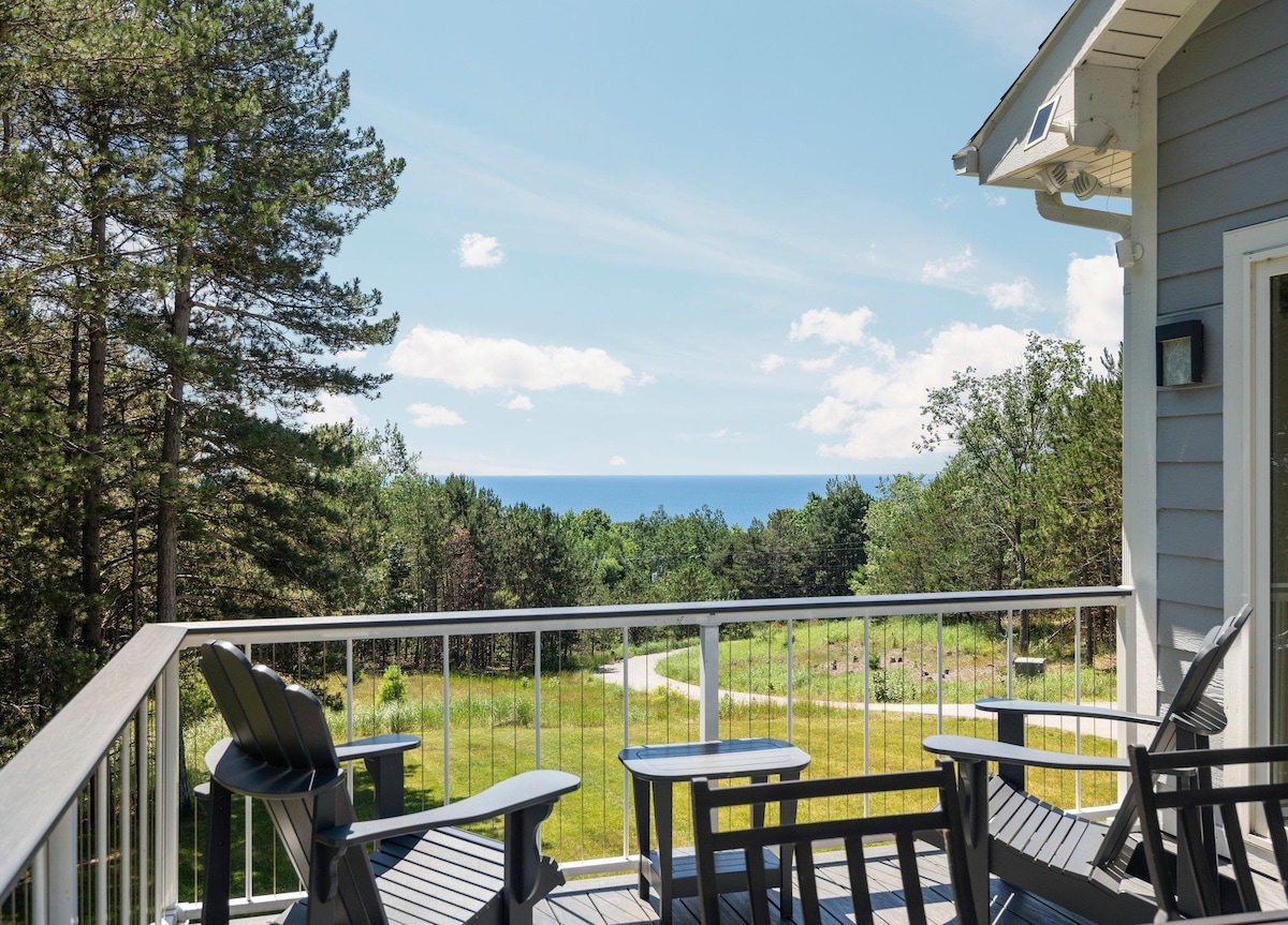 An outdoor deck features several Adirondack chairs arranged to face a panoramic view of lush greenery and distant Lake Michigan. The bright blue sky is dotted with soft white clouds, creating a serene backdrop for relaxation.