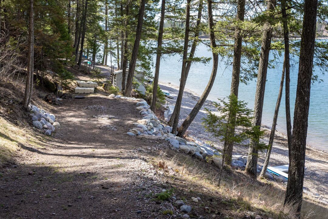 A gravel path lined with smooth stones leads towards a serene waterfront. Tall trees are positioned along the path, providing shade, while the clear water of Echo Lake reflects the shoreline. The beach area features a combination of pebbles and sand, creating a natural embankment.