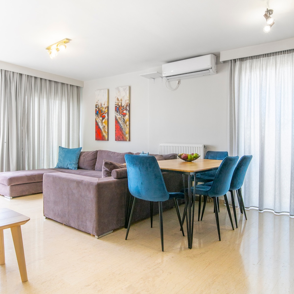 A spacious living area features a large sectional couch in a muted tone paired with blue accent chairs. A dining table is set adjacent, offering seating for multiple guests. Natural light filters through sheer curtains, enhancing the inviting environment.