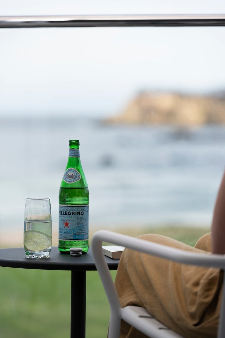 A green glass bottle of sparkling water is placed on a small, round table, accompanied by a tall glass filled with water and a slice of lime. The backdrop features a blurred view of the ocean and coastline, creating a serene atmosphere.