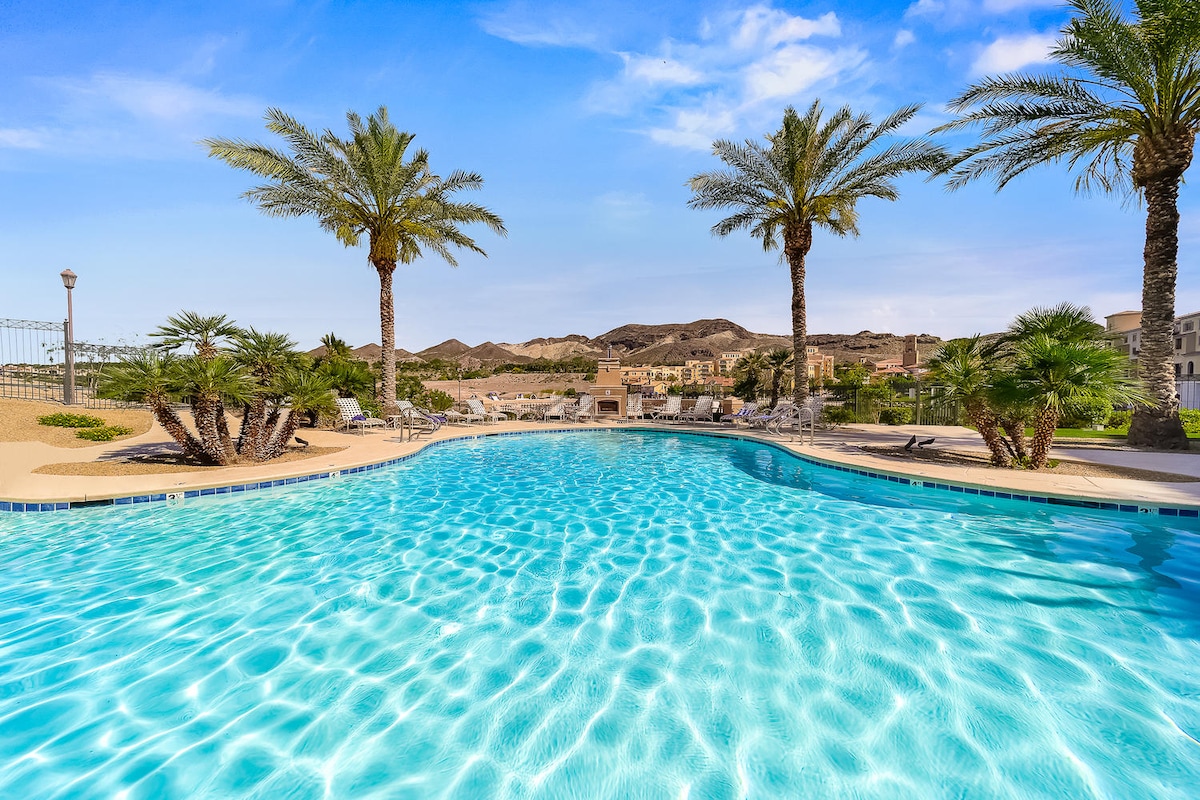 A clear, heated pool is surrounded by palm trees and lush greenery. Comfortable lounge chairs are positioned around the pool area, providing ample space for relaxation. The backdrop features gently rolling hills under a bright blue sky, creating a serene outdoor environment.