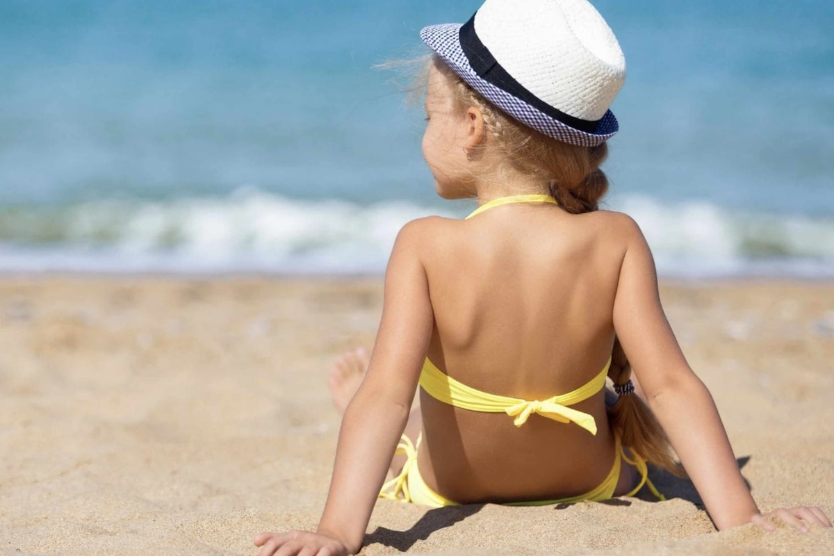 A child sits on the sandy beach, facing the calm blue sea. Wearing a yellow swimsuit and a straw hat, the child appears relaxed, with the gentle waves visible in the background. The warm sun creates a tranquil beach atmosphere.