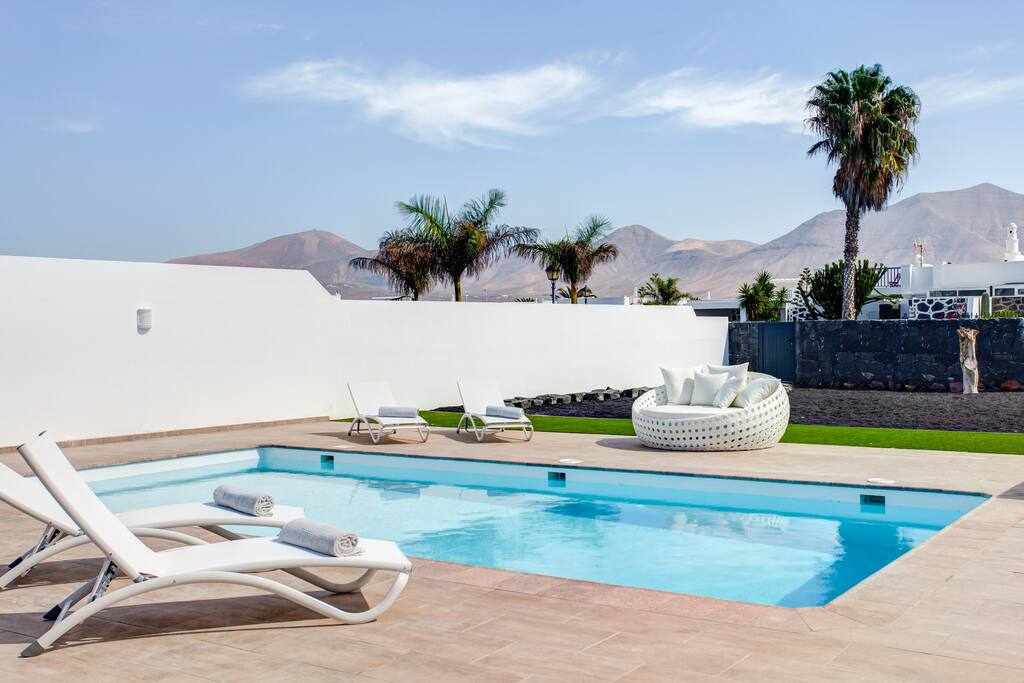The image displays a private swimming pool surrounded by a tiled patio. Two loungers are positioned beside the pool, while a round seating area with cushions is visible. Lush greenery and mountains create a serene backdrop, enhancing the tranquil setting.