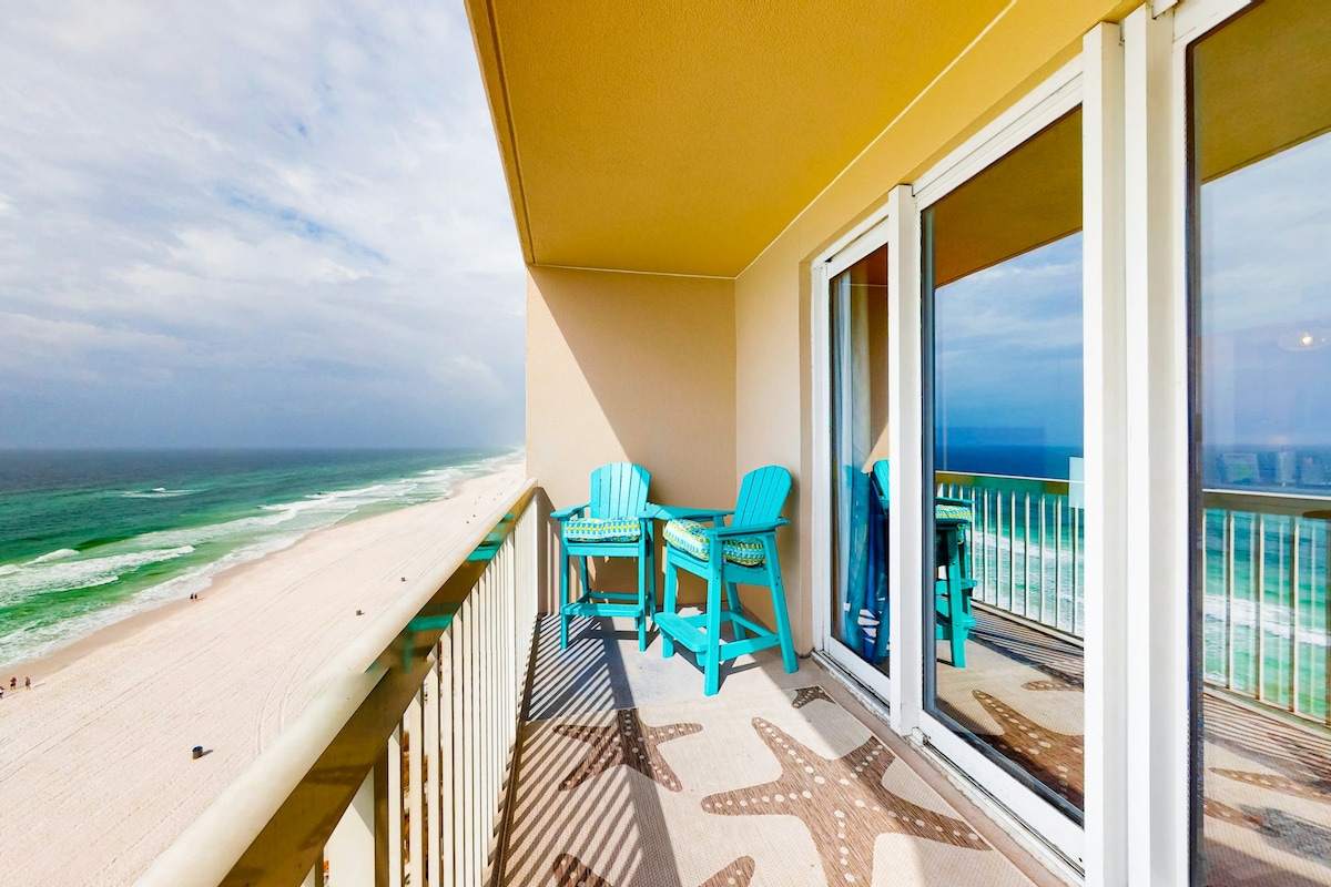 A covered balcony offers views of the emerald green waters and white sands of the Gulf of Mexico. Two turquoise chairs are positioned on the balcony's tiled surface, which features starfish designs. Large glass doors provide access to the interior living space while reflecting the coastal scenery.