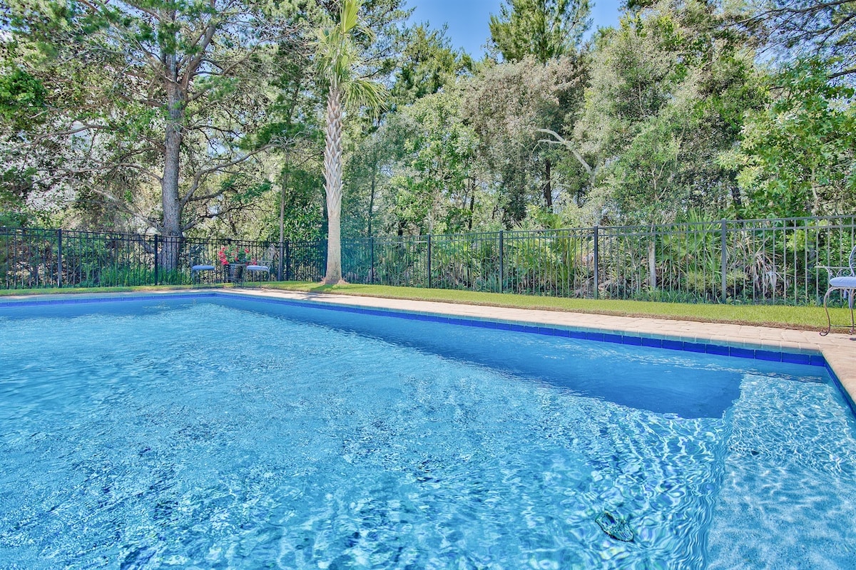 A private heated pool is surrounded by a secure fence and lush greenery, offering seclusion. The pool's clear blue water reflects the sunlight, and the landscape features palm trees and various plants, creating a tranquil outdoor setting.
