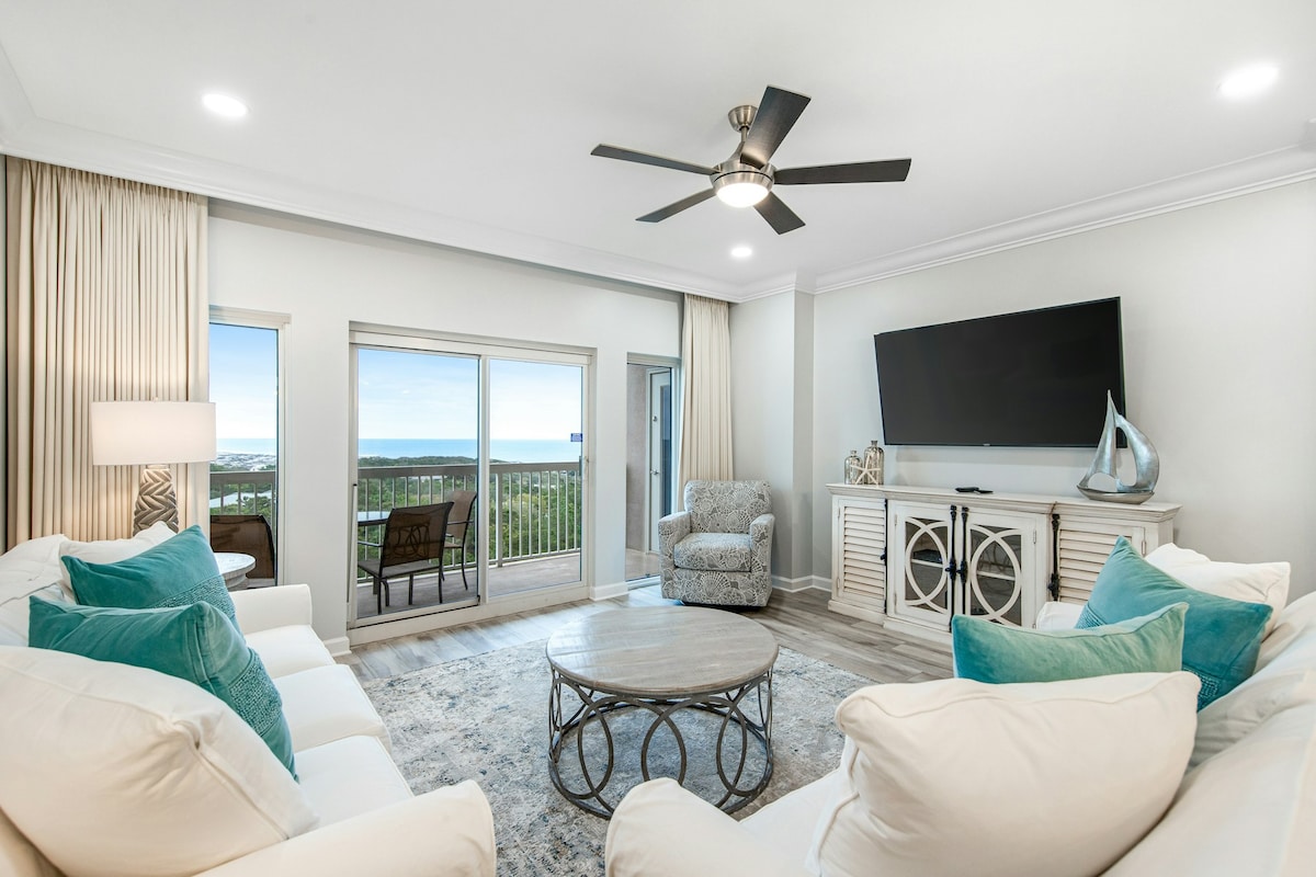 A spacious living area features pale walls and soft neutral furnishings. Large windows allow natural light to fill the space, revealing views of the surrounding landscape. A circular coffee table sits in the center, complemented by decorative blue cushions on the sofas.